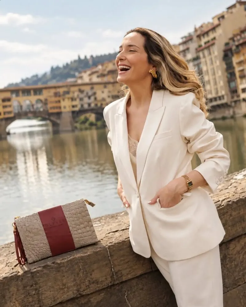 Mulher sorrindo de terno claro às margens do rio, com nécessaire vinho